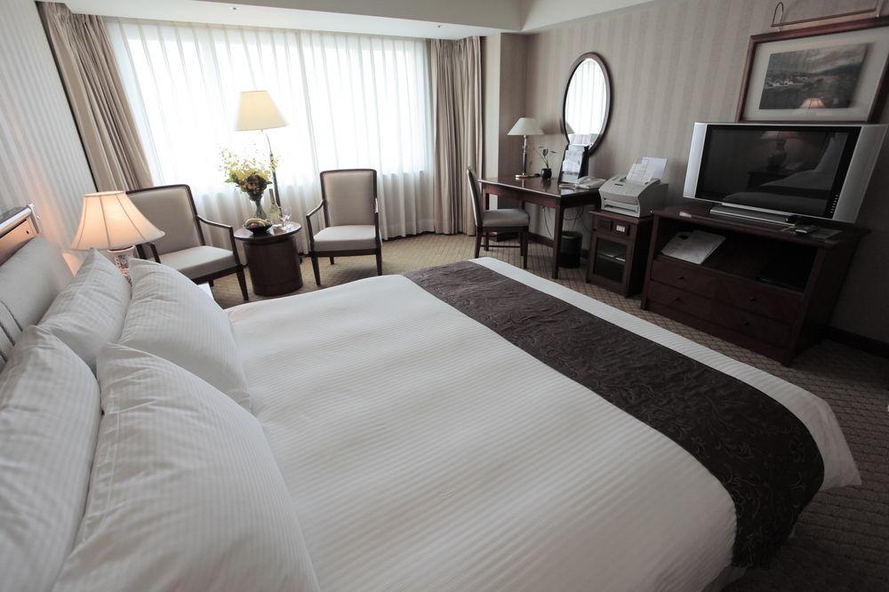 Evergreen Laurel Hotel - Taichung Esterno foto The photo shows a well-furnished hotel room. In the foreground, there is a large bed with white bedding and a dark-colored throw at the foot. Beside the bed, there is a bedside lamp. 

In the background, natural light filters through large windows co