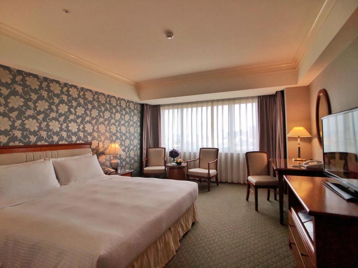 Evergreen Laurel Hotel - Taichung Esterno foto The photo shows a well-appointed hotel room. It features a large bed with white bedding and a decorative headboard. The walls are adorned with a patterned wallpaper, possibly floral, adding a touch of elegance. There is a window with sheer curtains t