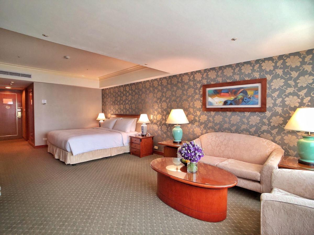 Evergreen Laurel Hotel - Taichung Esterno foto The photo shows a hotel room that features a well-furnished space. It includes a large bed with a light-colored bedspread, a comfortable sofa, and armchair. There's a coffee table with a vase of flowers in the center. The room has a decorative wallpa