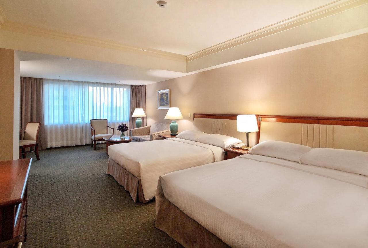 Evergreen Laurel Hotel - Taichung Esterno foto The photo shows a hotel room featuring two queen-sized beds with white linens. The room is well-lit and has a warm, neutral color palette. There are bedside lamps on each side of the beds and a small table. To the side, there's a comfortable armchair