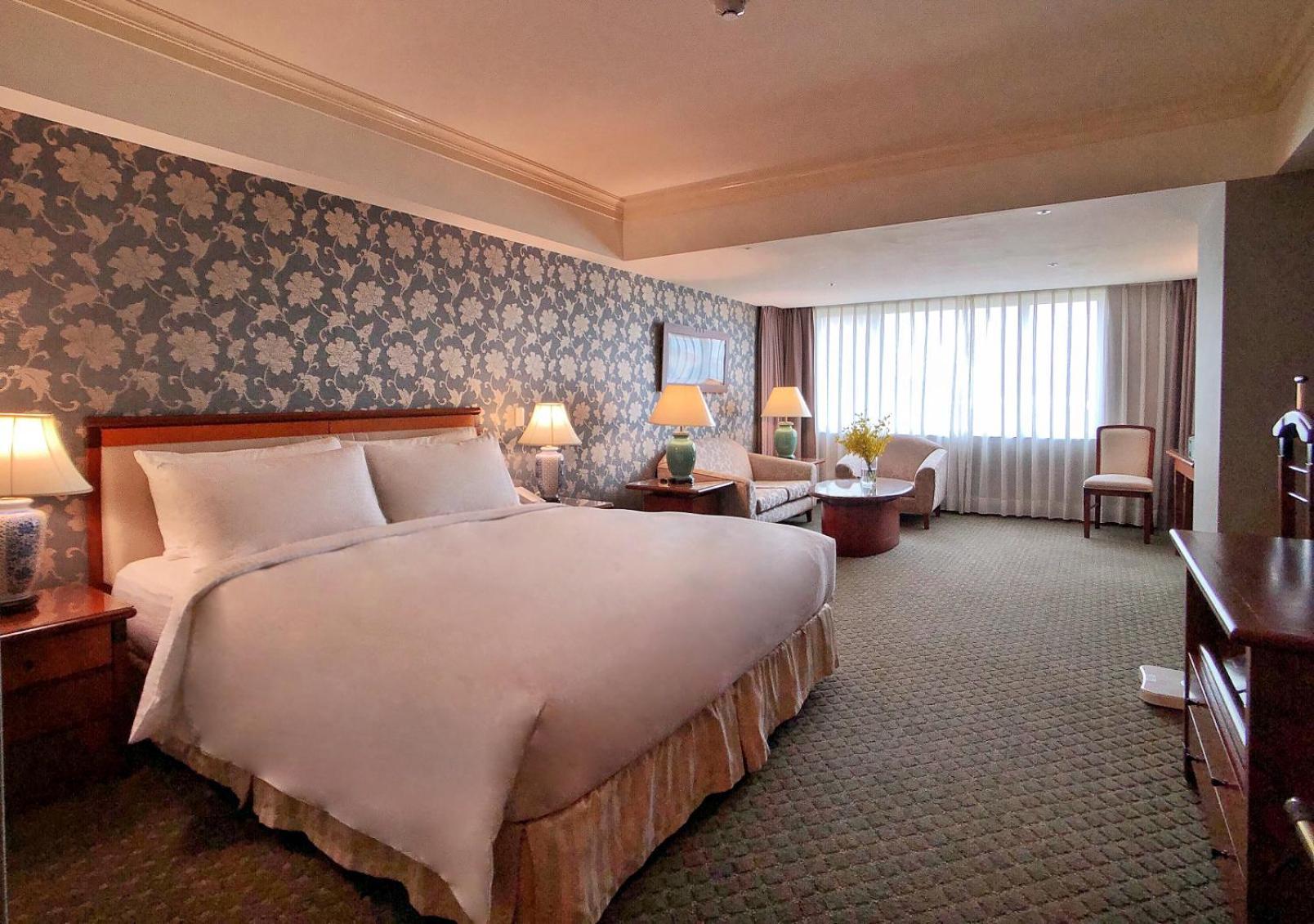 Evergreen Laurel Hotel - Taichung Esterno foto The photo shows a hotel room with a spacious and comfortable layout. It features a large bed with white bedding, placed against a decorative wallpapered wall. On either side of the bed, there are lamps providing soft lighting. In addition to the bed,