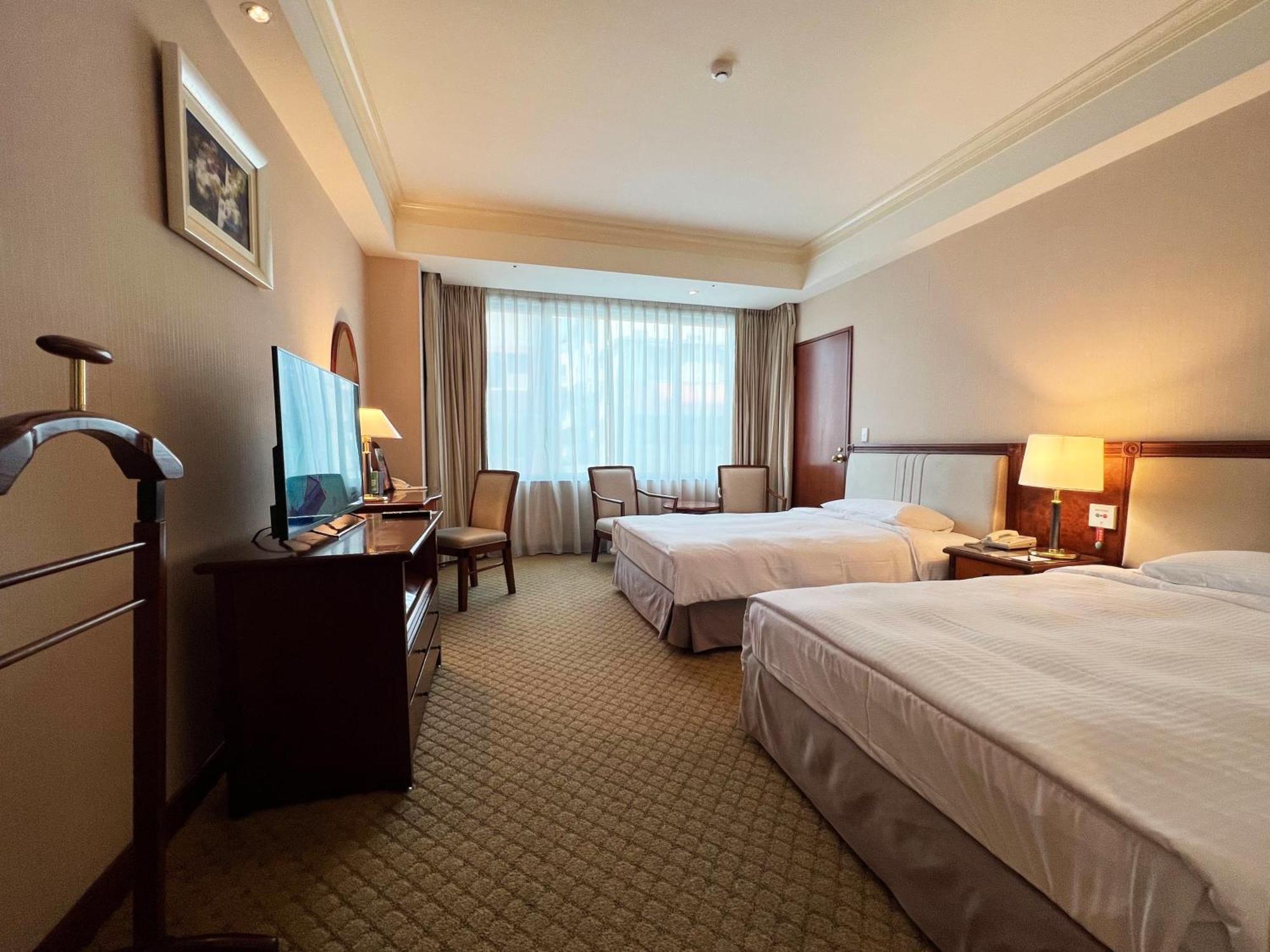 Evergreen Laurel Hotel - Taichung Esterno foto The photo shows a hotel room featuring two twin beds placed parallel to each other. The room has a warm color palette and includes a large window with sheer curtains allowing natural light to illuminate the space. There is a wooden desk and two chair