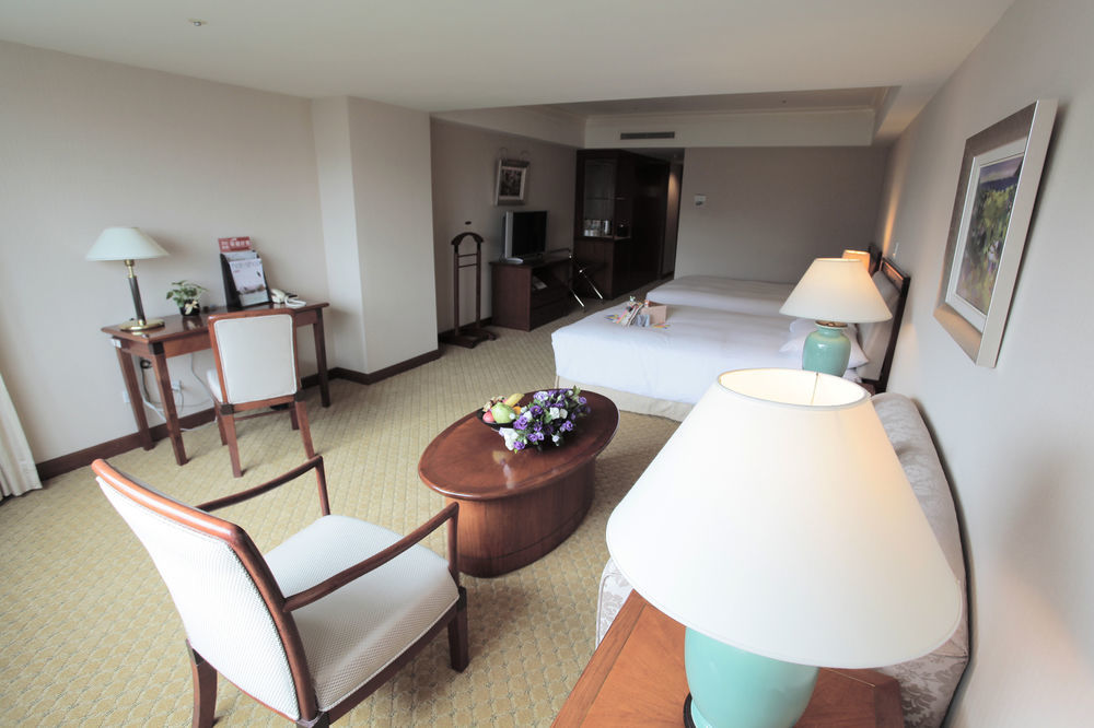 Evergreen Laurel Hotel - Taichung Esterno foto The photo shows an interior view of a hotel room. It features two beds, a coffee table with a small decorative arrangement of flowers, and a comfortable chair. There's a desk and a lamp, along with another larger lamp on the table. In the background,