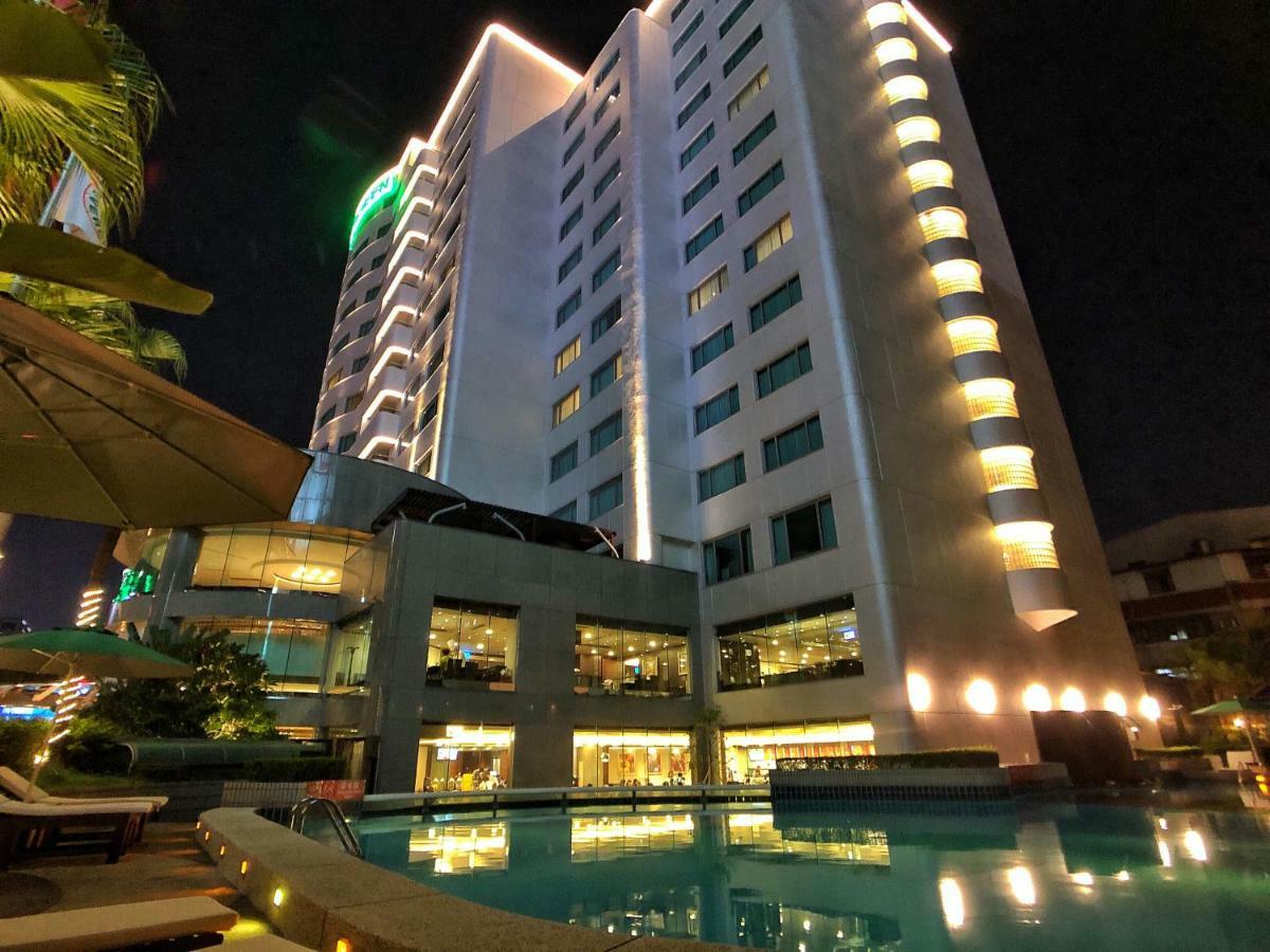 Evergreen Laurel Hotel - Taichung Esterno foto The photo shows a tall hotel building at night, illuminated by lights. The architecture appears modern, with several floors and large windows. In the foreground, there is a swimming pool that reflects the lights from the building, creating a serene a