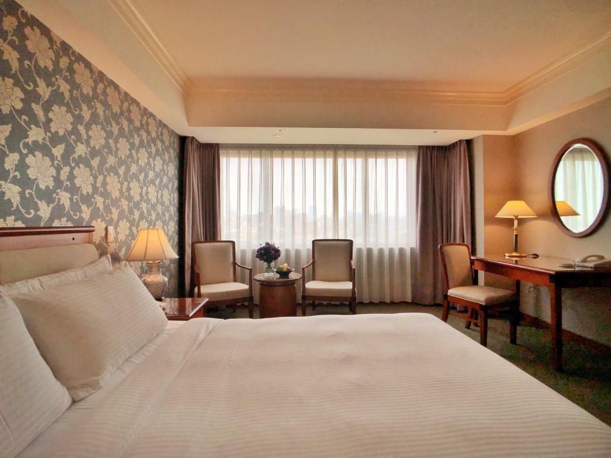 Evergreen Laurel Hotel - Taichung Esterno foto The photo shows a hotel room with a comfortable and inviting design. There is a large bed with crisp white bedding on the left side of the image. To the right, there are two elegant chairs with a small round table between them, adorned with a bouquet