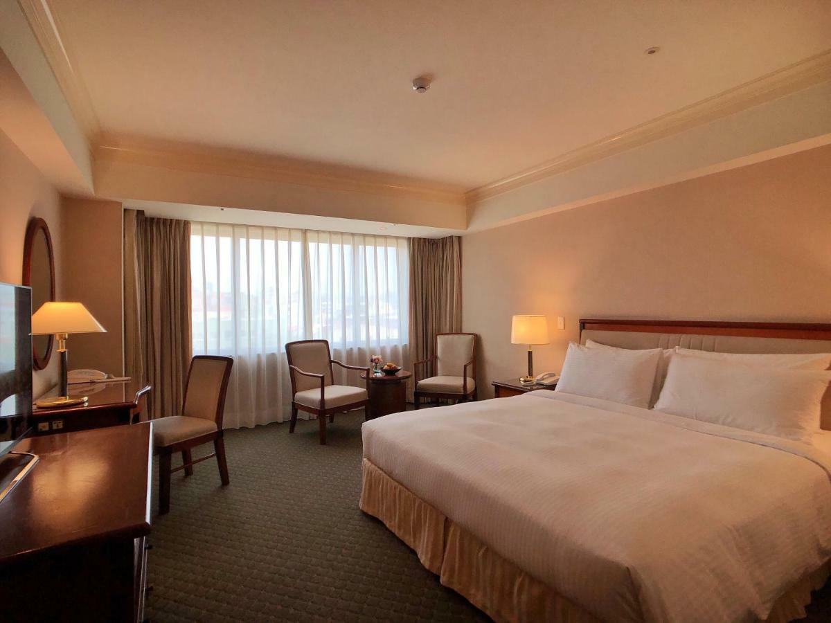 Evergreen Laurel Hotel - Taichung Esterno foto The photo shows a hotel room featuring a large bed with white linens, a wooden bedside table with a lamp, and a flat-screen television. There are two chairs with a small table next to them positioned by a large window, which has sheer curtains lettin