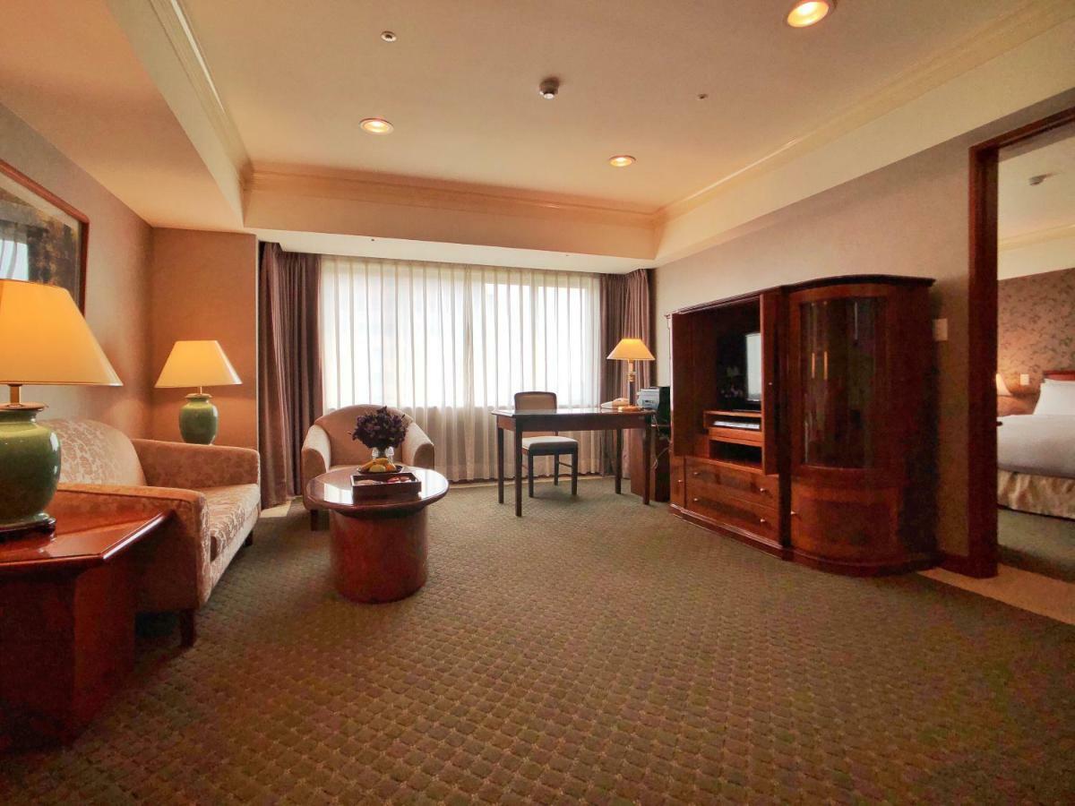 Evergreen Laurel Hotel - Taichung Esterno foto The photo shows a cozy hotel suite or living area. There is a sofa and an armchair arranged around a small coffee table. A desk with a chair is positioned near a large window covered with sheer curtains that allow natural light to enter the room. Two