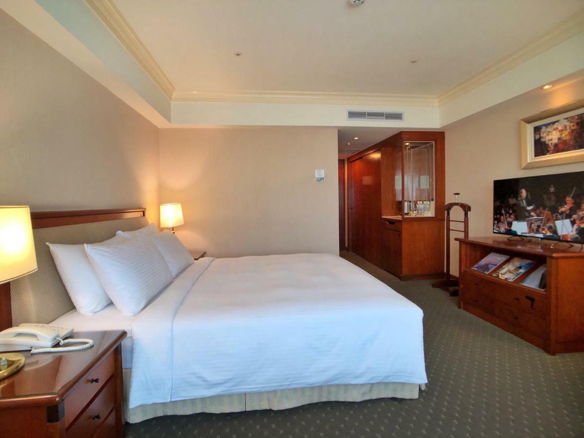 Evergreen Laurel Hotel - Taichung Esterno foto The photo shows a hotel room featuring a comfortable bed with white linens and pillows, positioned against a light-colored wall. There is a wooden bedside table with a lamp and an open book next to the bed. On the opposite side, there’s a large flat-