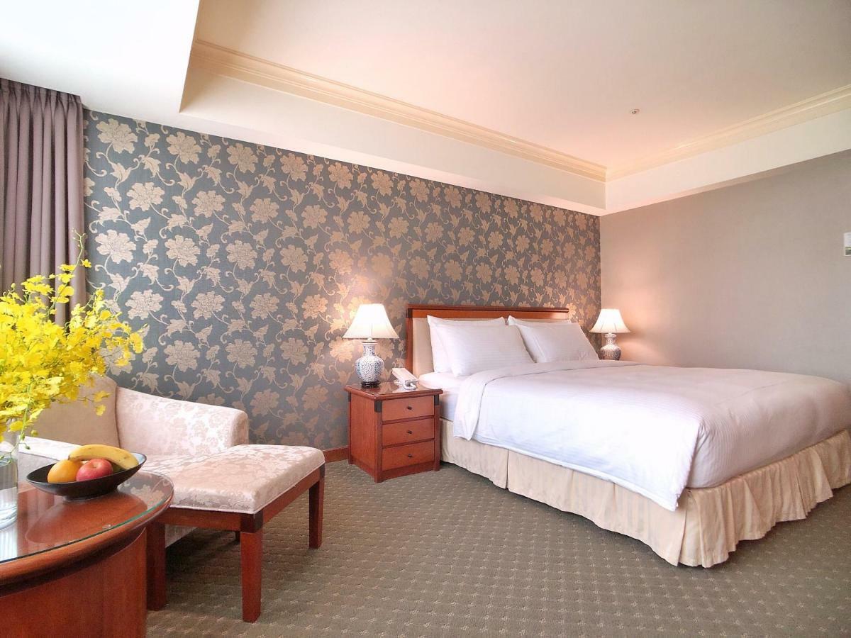 Evergreen Laurel Hotel - Taichung Esterno foto The photo shows a well-decorated hotel room. It features a large bed with white bedding, situated against a patterned wall with floral designs. On either side of the bed are bedside tables with lamps. There is a small seating area that includes a cha
