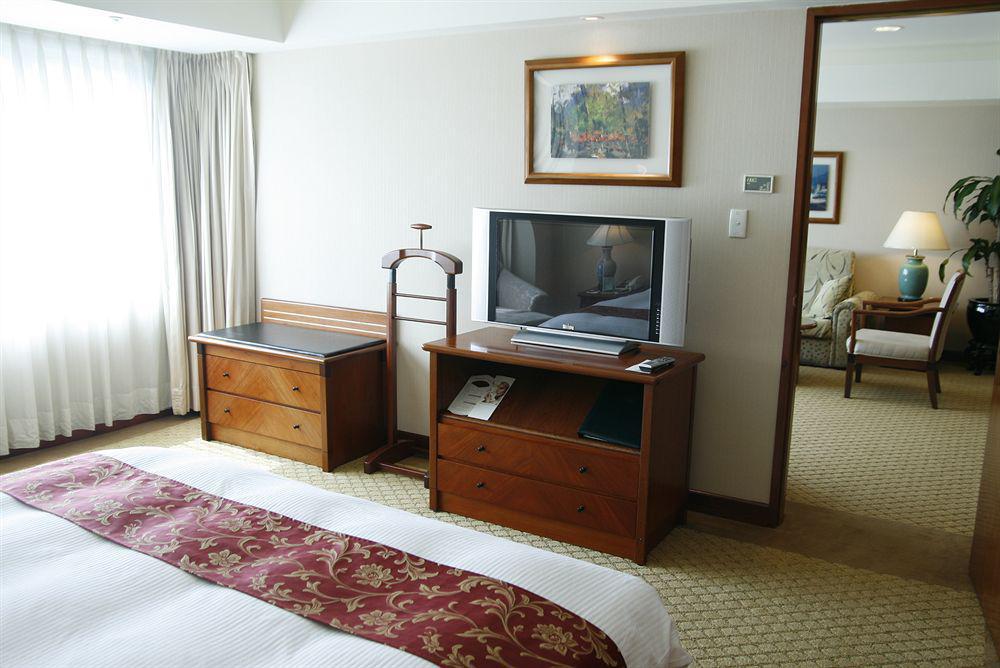 Evergreen Laurel Hotel - Taichung Esterno foto The photo depicts a hotel room interior. In the foreground, there is a neatly made bed with a decorative red bedspread. To the left, a large window allows natural light to fill the room, with light-colored curtains drawn to one side. 

Next to the be