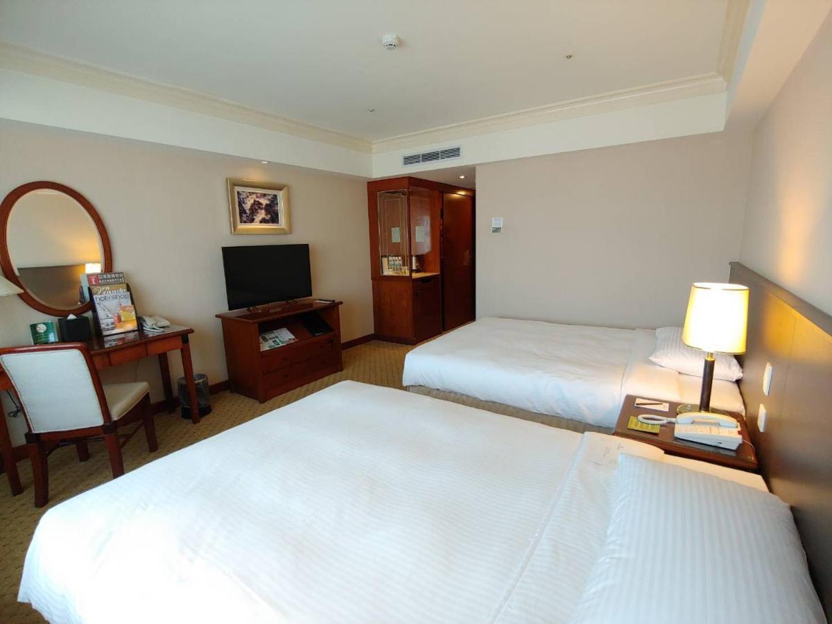 Evergreen Laurel Hotel - Taichung Esterno foto The photo shows a hotel room with two beds, neatly made with white linens. There is a desk and chair in one corner, accompanied by a flat-screen TV mounted on the wall. A large mirror and a wooden cabinet are also visible. The room is illuminated by 