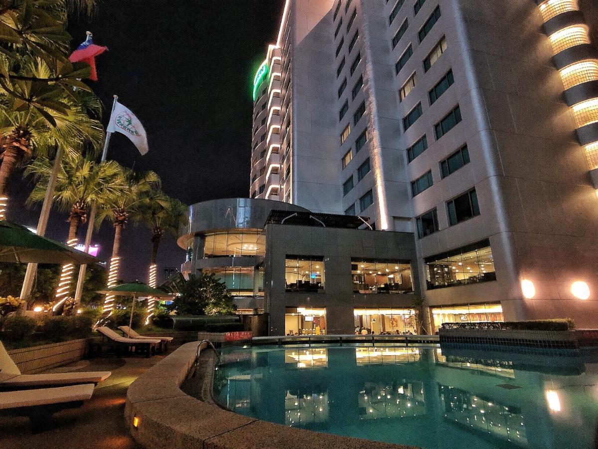 Evergreen Laurel Hotel - Taichung Esterno foto The photo depicts a modern hotel or building at night, featuring a sleek facade illuminated by lights. In the foreground, there is a swimming pool that reflects the surrounding lights and palm trees. There are lounge chairs near the pool, and the atm
